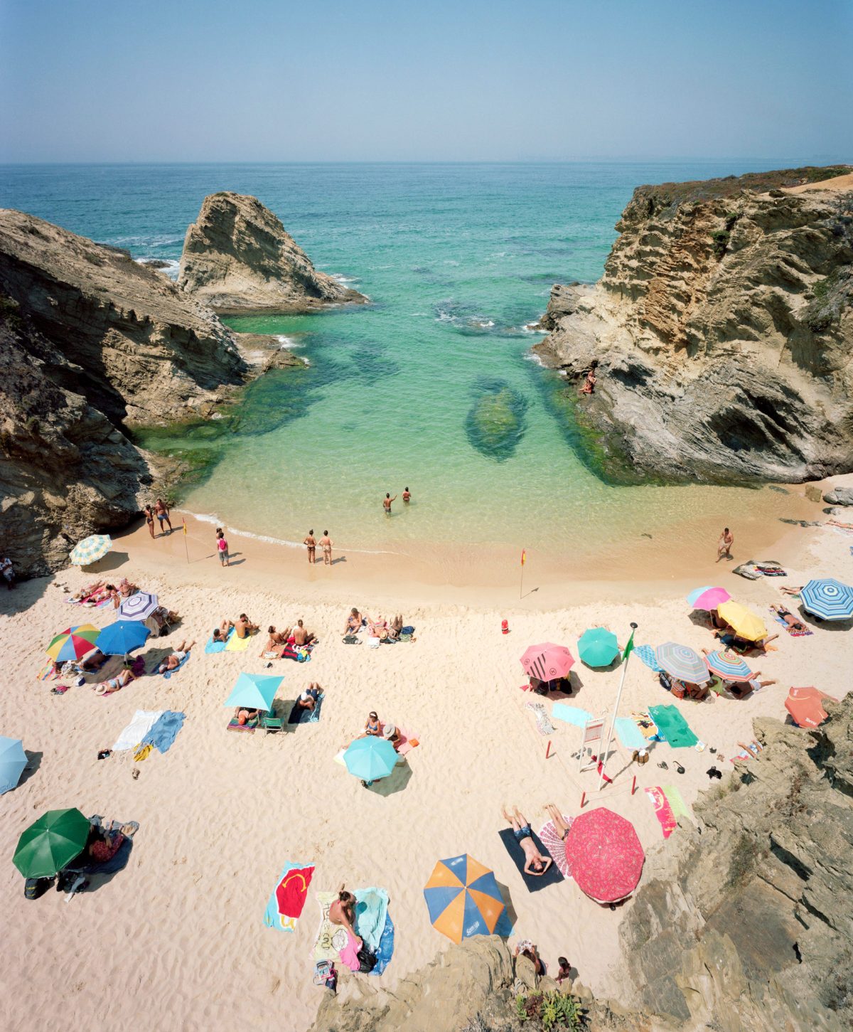 Aerial view of a secluded cove beach framed by dramatic limestone cliffs, with colorful umbrellas and beachgoers scattered across golden sand and turquoise shallow waters.