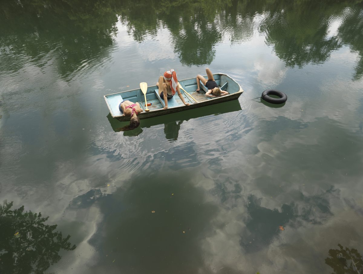 A small rowboat floats on still water surrounded by green trees and their reflections. Three figures recline in the boat amid oars and supplies, while a dark tire bobs nearby in the calm, mirror-like surface.