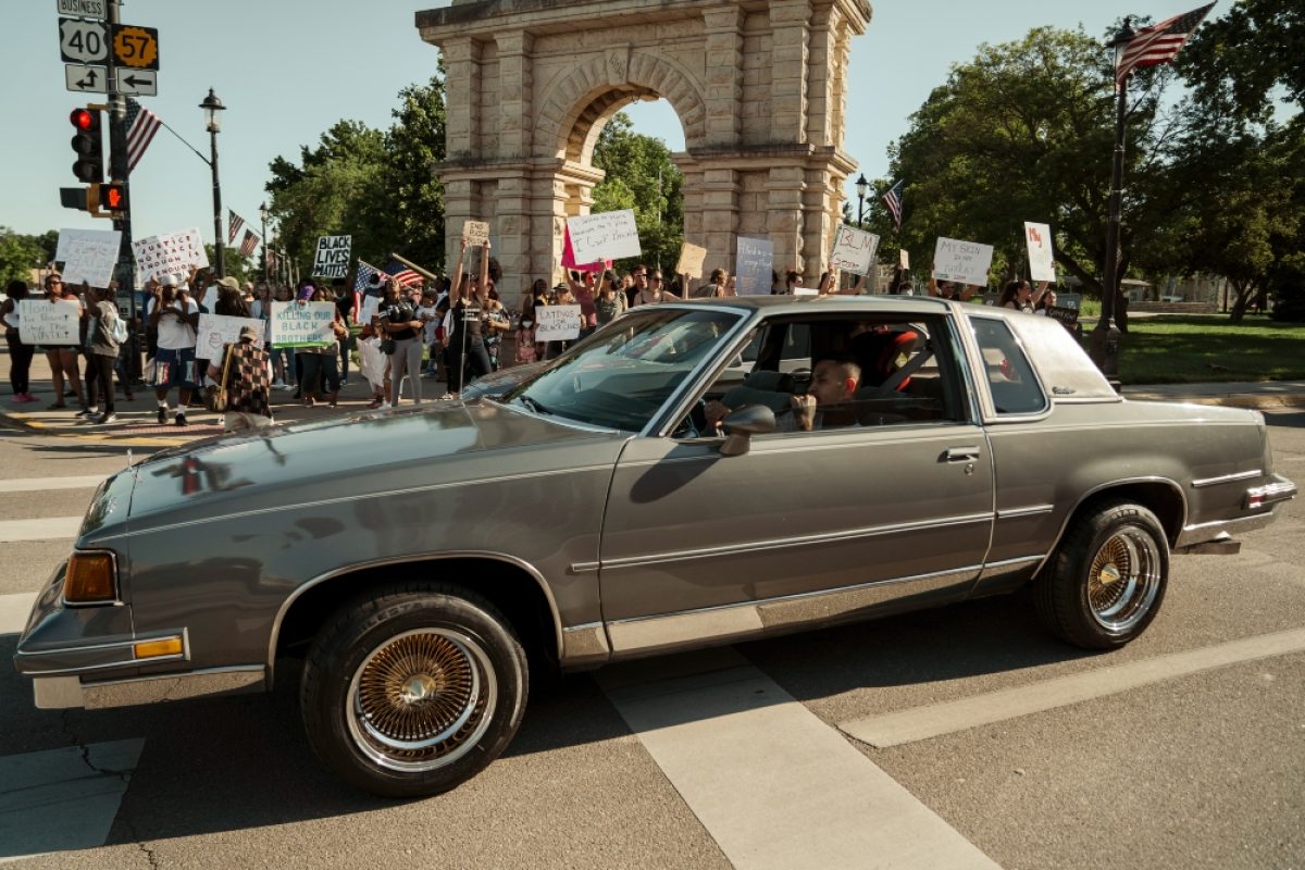 A gray sedan with gold-trimmed wheels drives through a city street lined with protesters holding signs. Behind the vehicle, a large stone arch monument is visible, with American flags and crowds gathered in the background under clear sky.