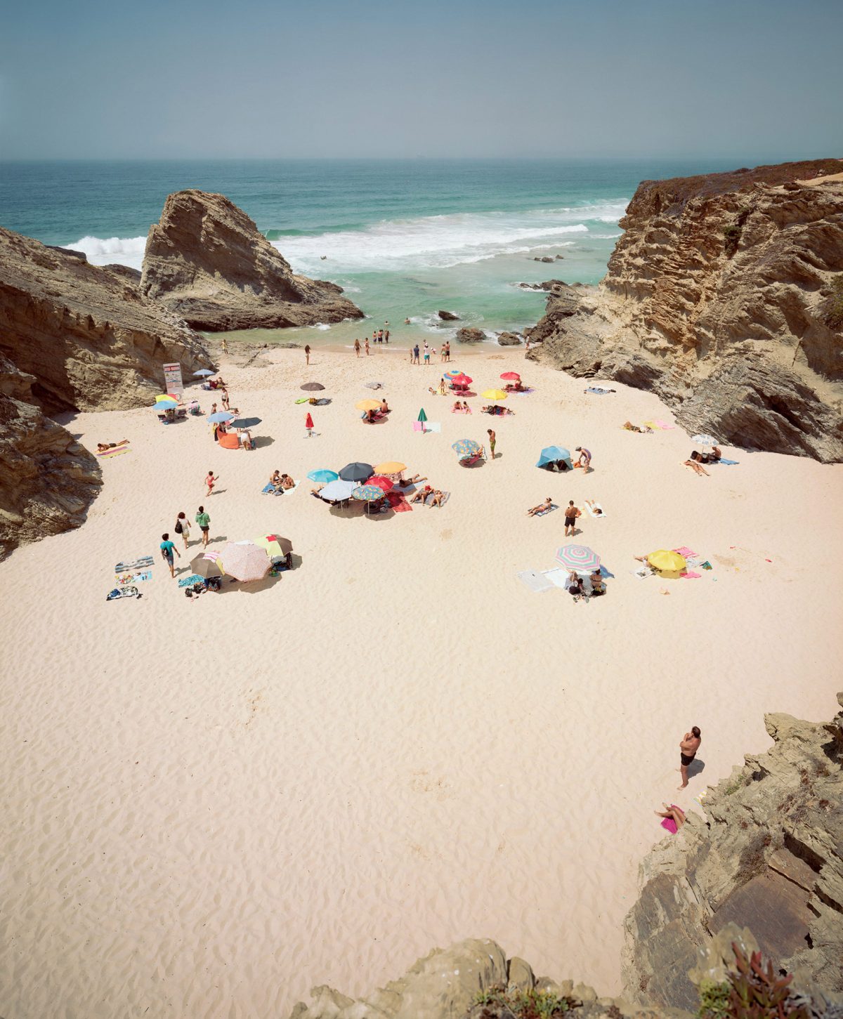 Aerial view of a sandy cove enclosed by dramatic rocky cliffs, with numerous beachgoers scattered across the sand under colorful umbrellas and beach tents, the turquoise ocean and white-capped waves visible beyond.