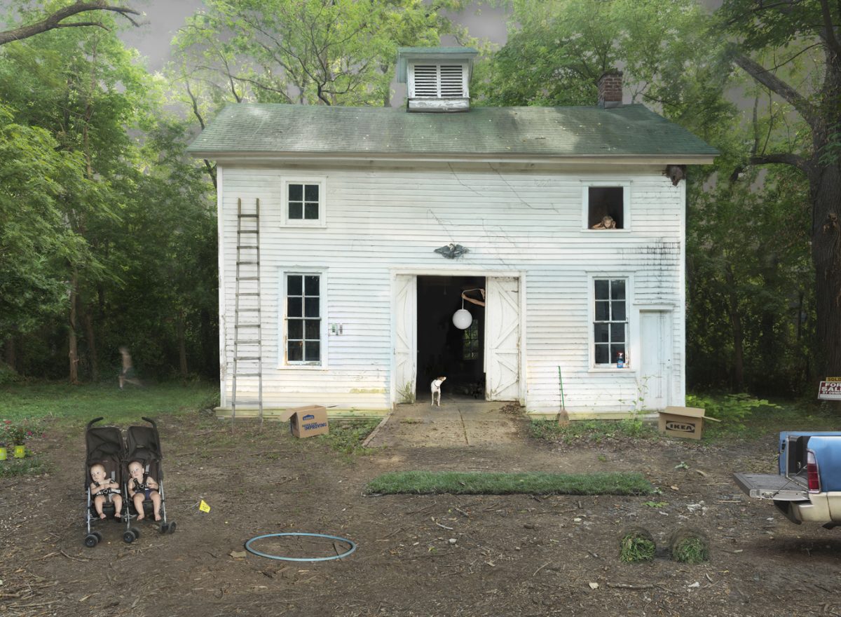 A white wooden house surrounded by dense green trees sits in a dirt yard scattered with children's toys, including a double stroller on the left, a hula hoop, and cardboard boxes. Figures appear in several windows and doorways of the modest two-story structure, while a small dog stands near the open front entrance. The scene is composed as a centered, frontal view with a documentary-like quality.