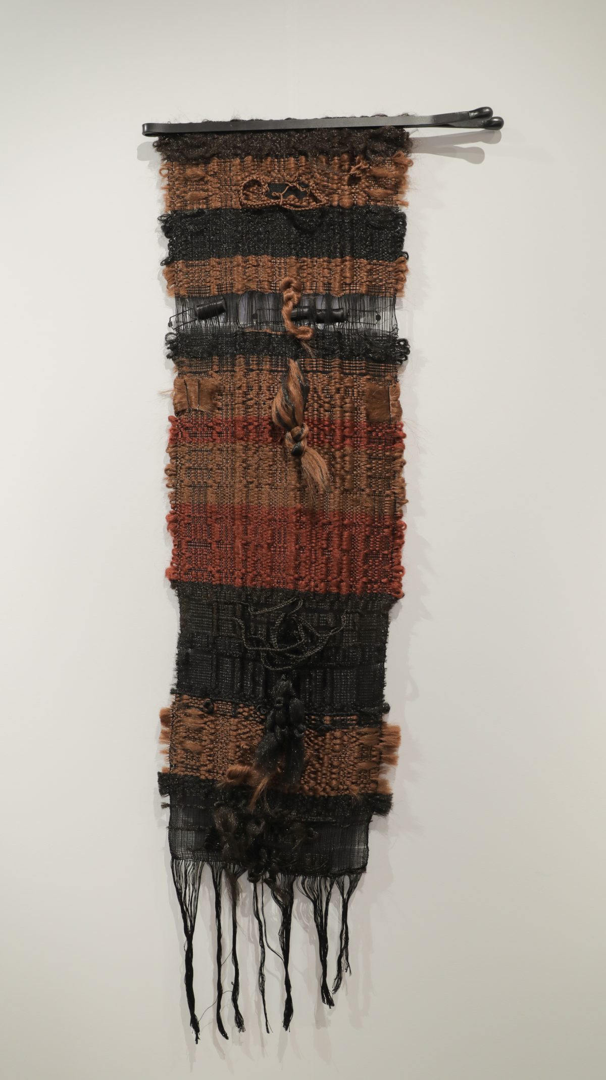 Vertical textile wall hanging composed of alternating bands of woven fabric in copper, rust, and black tones, with synthetic hair woven throughout and secured with satin rollers and bobby pins at intervals. Black fringe extends from the bottom edge.