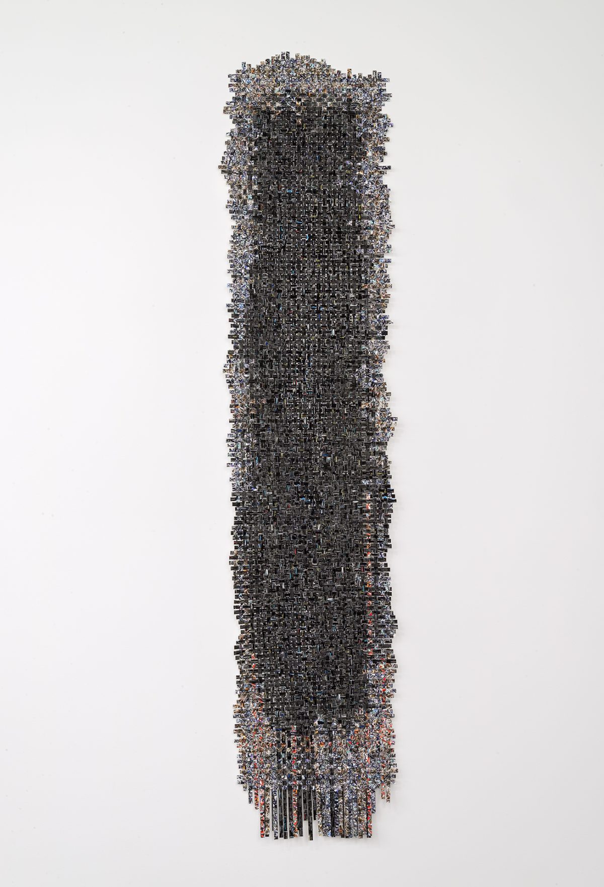 A tall, narrow vertical composition densely composed of hundreds of small rectangular elements in dark grays, blacks, and muted earth tones, arranged in a tightly woven grid pattern. The form tapers slightly at top and bottom, with loose strands or threads extending downward, creating a textile-like appearance against a white background.