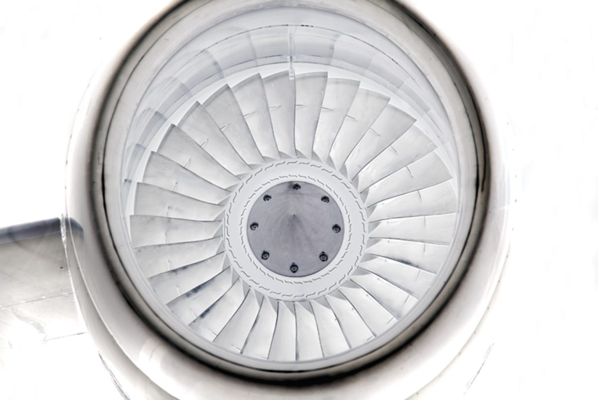Overhead view of a jet engine's fan blades radiating outward in concentric white metal rings, photographed from directly above. The geometric pattern creates a symmetrical spiral composition with a central circular hub, rendered in grayscale tones.