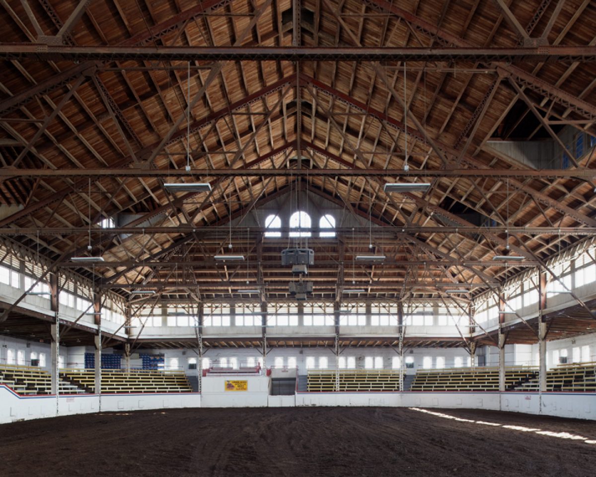 Interior view of a vast wooden-beamed arena with a dirt floor, yellow and white bleachers lining the sides, and natural light streaming through arched windows and skylights in the peaked roof structure.