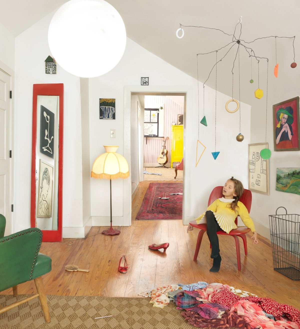 A young girl in a yellow sweater sits in a red chair gazing upward in a bright, eclectic interior space. The room features white walls, natural wood flooring, colorful artworks and objects including a large illuminated ceiling fixture, a hanging mobile with geometric shapes and ornaments, a yellow-fringed floor lamp, and scattered clothing on the floor, creating a whimsical, lived-in domestic scene filled with vibrant reds, greens, yellows, and blues.