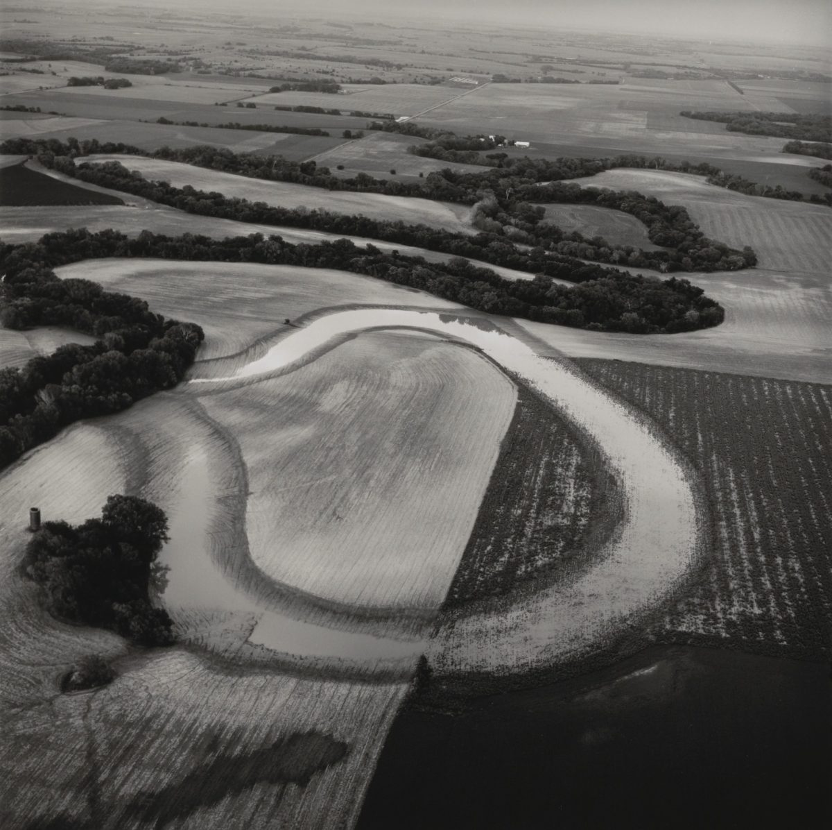 Aerial black-and-white photograph of a winding river creating an oxbow formation through agricultural farmland, with curved fields showing distinct plow patterns and tree-lined banks contrasting against lighter cultivated areas.
