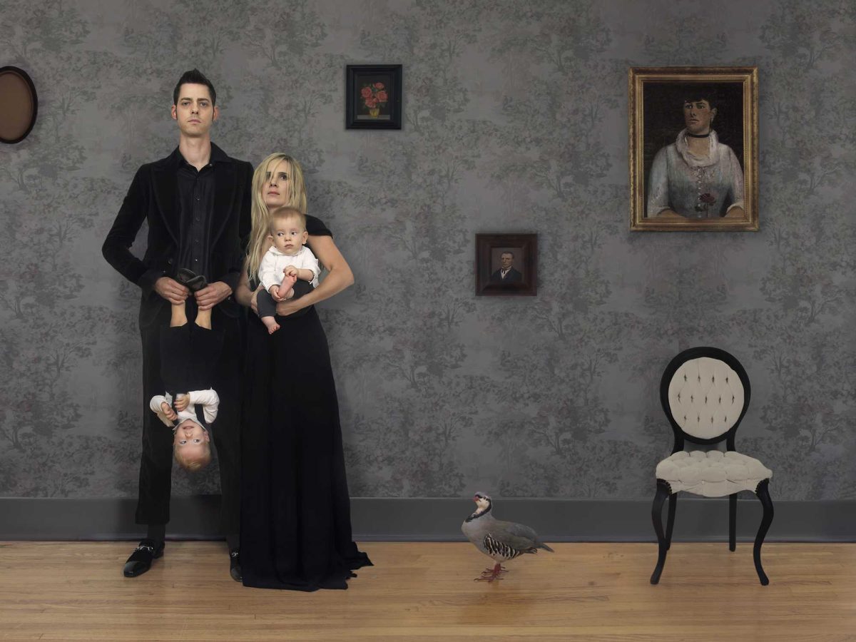 A man and woman in black clothing stand against a damask-patterned wall, with the woman holding two infants. A child hangs upside down from the man's arm, and a small goose walks across the wooden floor in the foreground. Period portraits and a round-backed chair are mounted on the wall behind them.