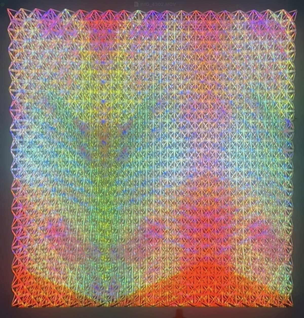 A large square composition of densely packed triangular forms creating an intricate lattice pattern. The work features a vivid color gradient transitioning from cool purples and blues in the upper left through vibrant greens in the center to warm oranges and reds in the lower right, with subtle iridescent and reflective qualities throughout.