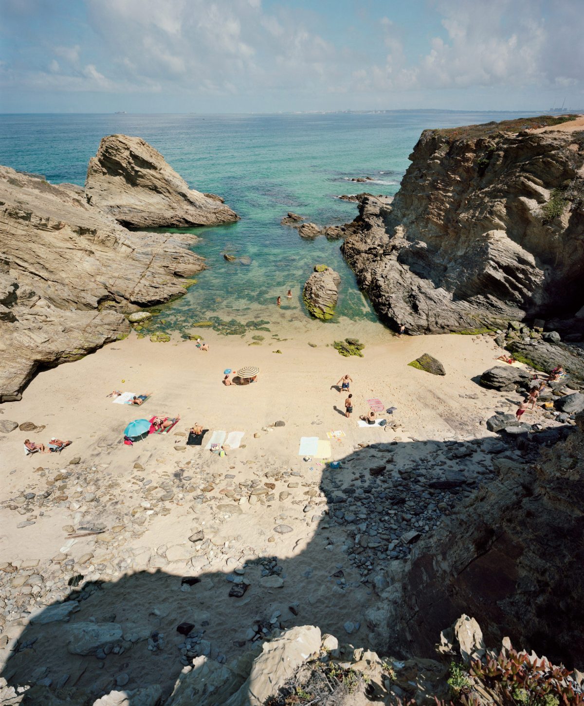 Aerial view of a secluded sandy beach nestled between dramatic rocky outcrops and turquoise water, with scattered beachgoers and colorful umbrellas dotting the pale sand below.