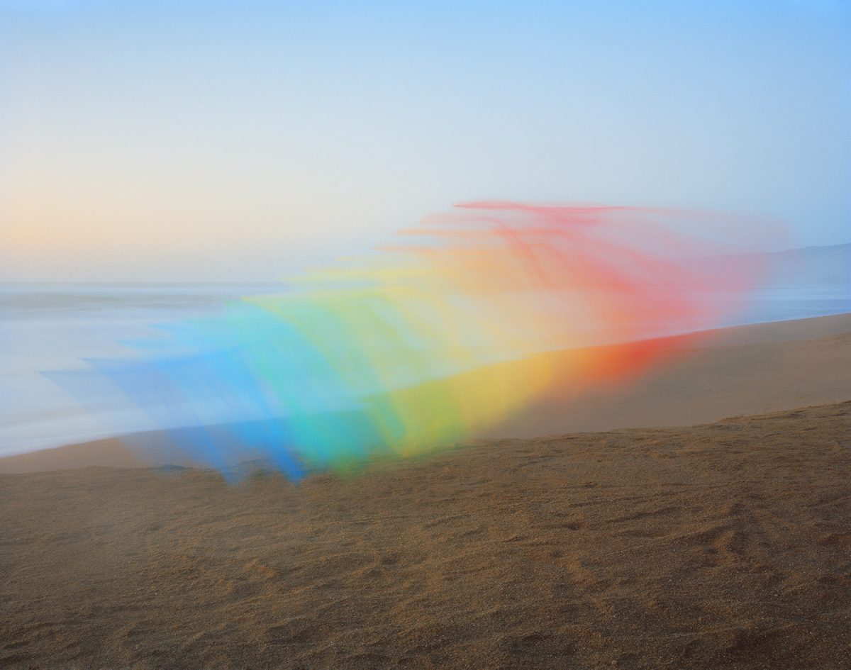 A rainbow gradient of tulle fabric stretches diagonally across a misty beach landscape, its translucent layers transitioning from blue through green, yellow, orange, and pink against a pale dawn sky and sandy shore.