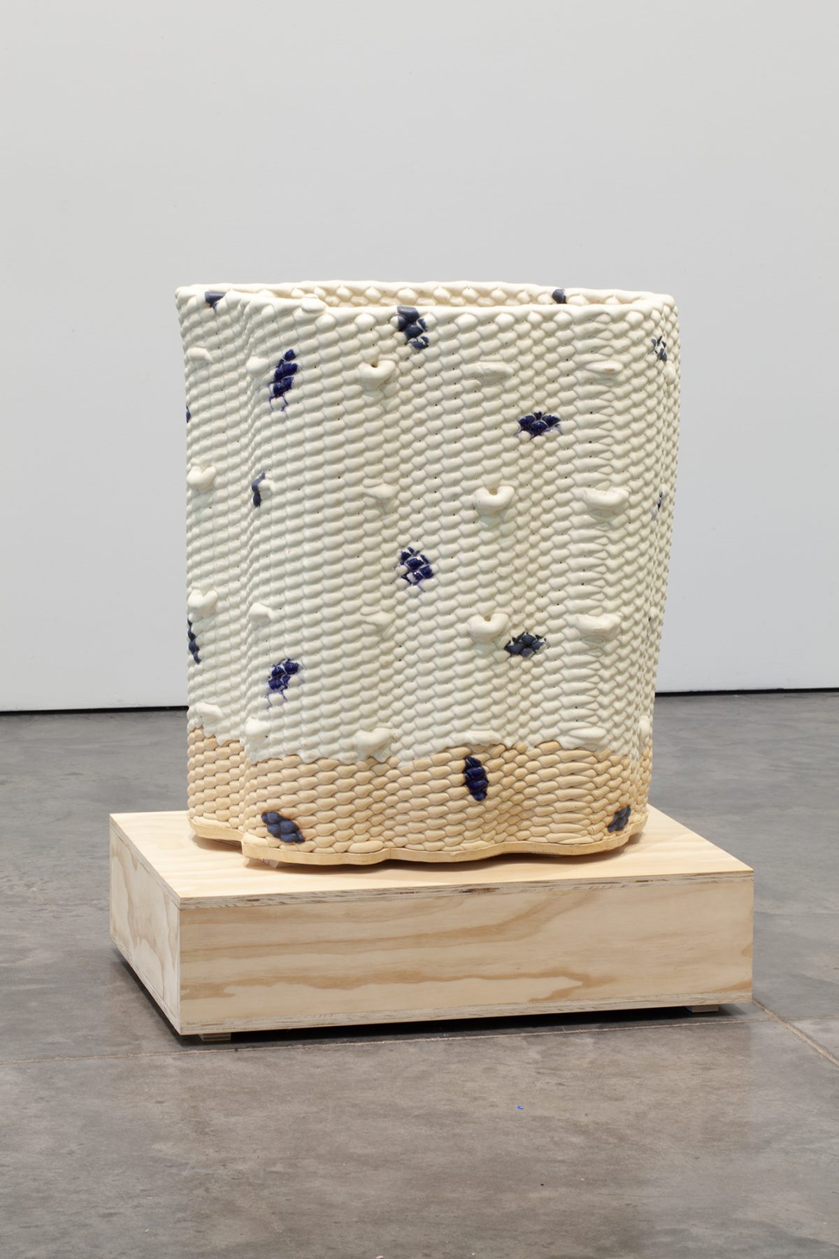 A cylindrical ceramic vessel with a textured, bumped surface in cream and pale yellow tones, scattered with dark blue glazed marks. The piece sits on a light wooden base against a gallery floor and white wall.