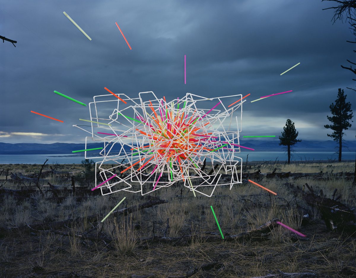 Neon-colored straws and white geometric wire frames arranged in a chaotic sculptural composition float above a desert landscape with a large lake visible in the distance under a dramatic cloudy sky.