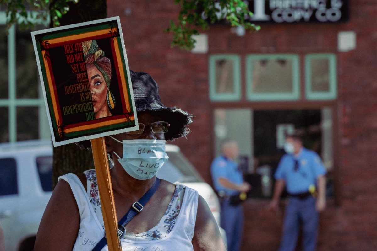 A protester wearing a face mask holds a wooden sign featuring a portrait of a woman's face in warm orange and red tones with text overlaid, standing before a brick building with uniformed officers visible in the background.