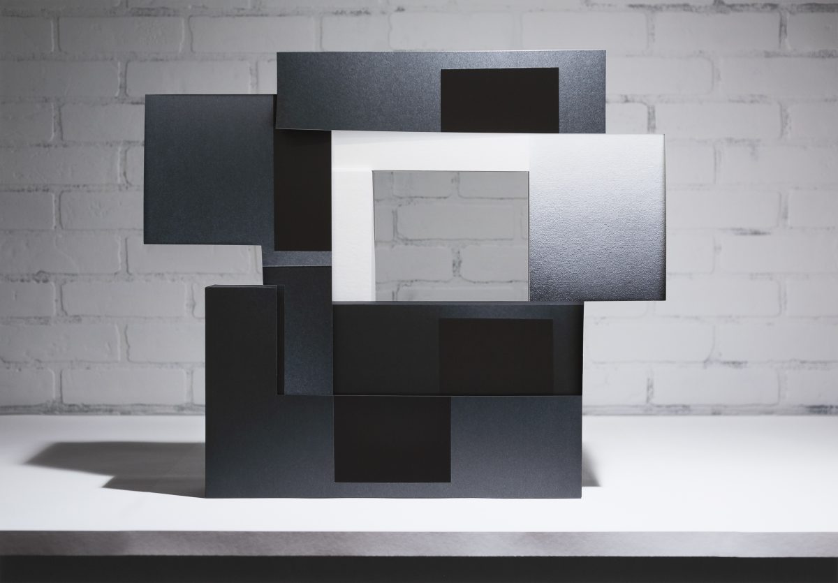 Layered rectangular panels in black, dark gray, and white arranged in an overlapping, three-dimensional composition against a white brick wall. The stacked forms create depth through varying planes and tones, with some surfaces appearing textured or printed, casting subtle shadows on the white surface beneath.