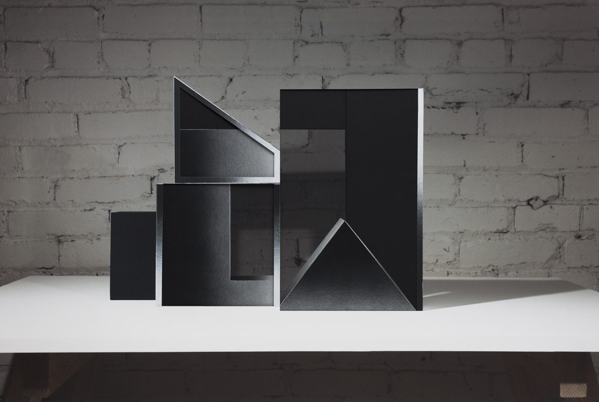 Abstract geometric sculpture made of black layered materials arranged in ascending rectangular and triangular forms on a white platform. The composition features sharp angles, cutouts, and overlapping planes that create dramatic shadows against a weathered brick wall.