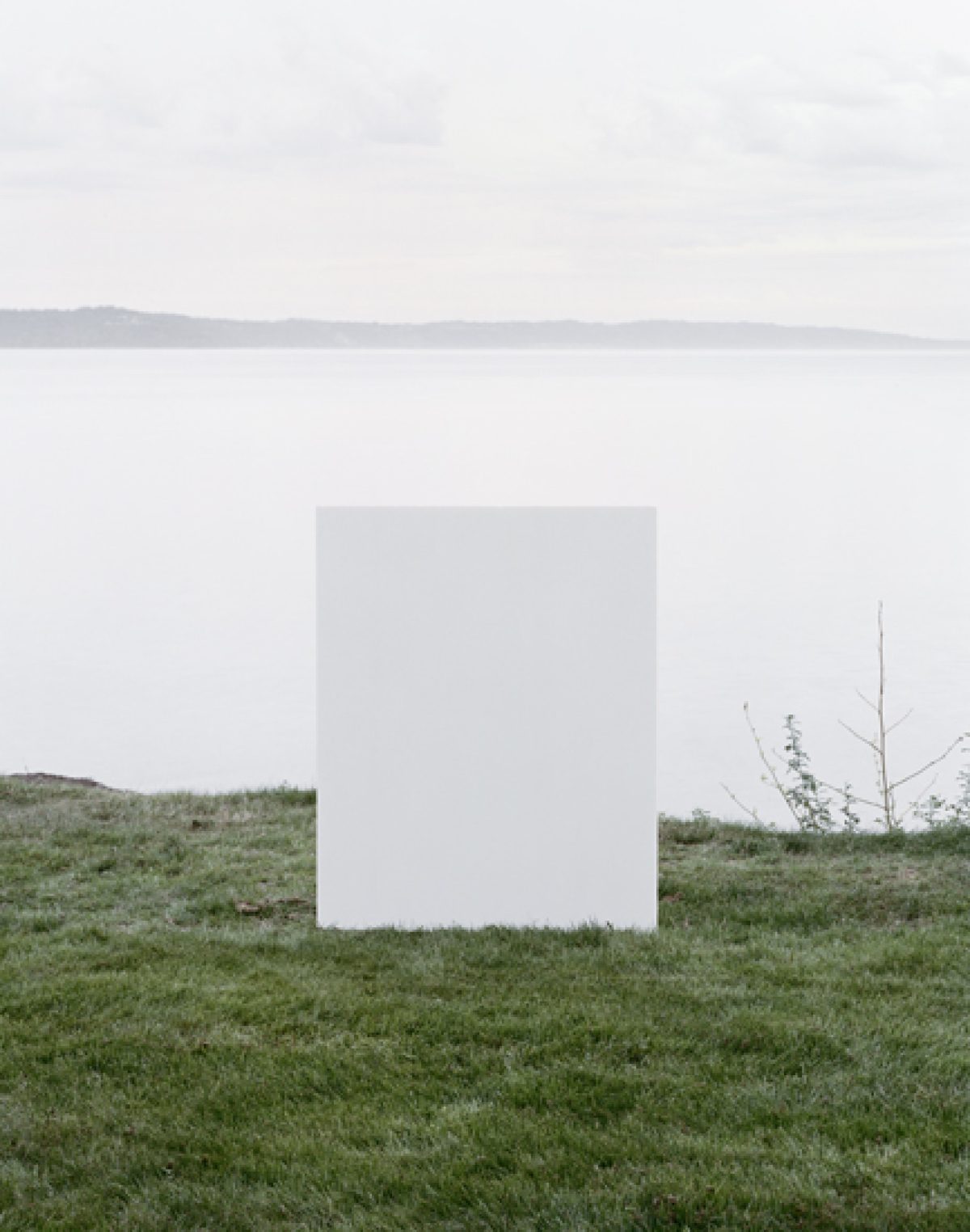 A large white rectangular form stands centered in a grassy field overlooking a calm body of water and distant shoreline under an overcast sky. Sparse dried vegetation and a leafless branch appear to the right of the geometric structure.