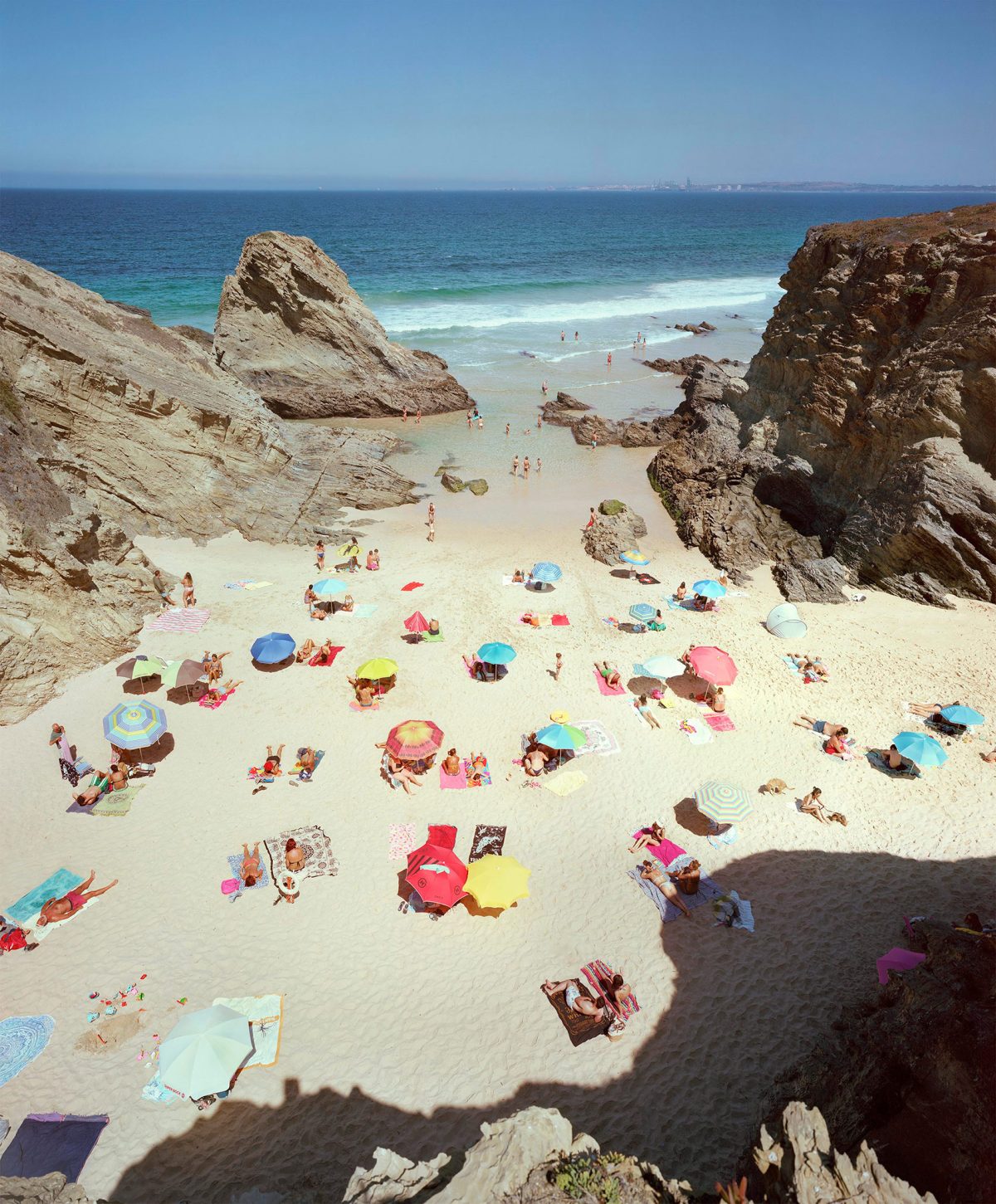 Aerial view of a crowded beach nestled between dramatic rocky outcrops, with numerous colorful umbrellas and towels dotting the pale sand. Turquoise and blue waters with gentle waves extend to the horizon under clear sky.