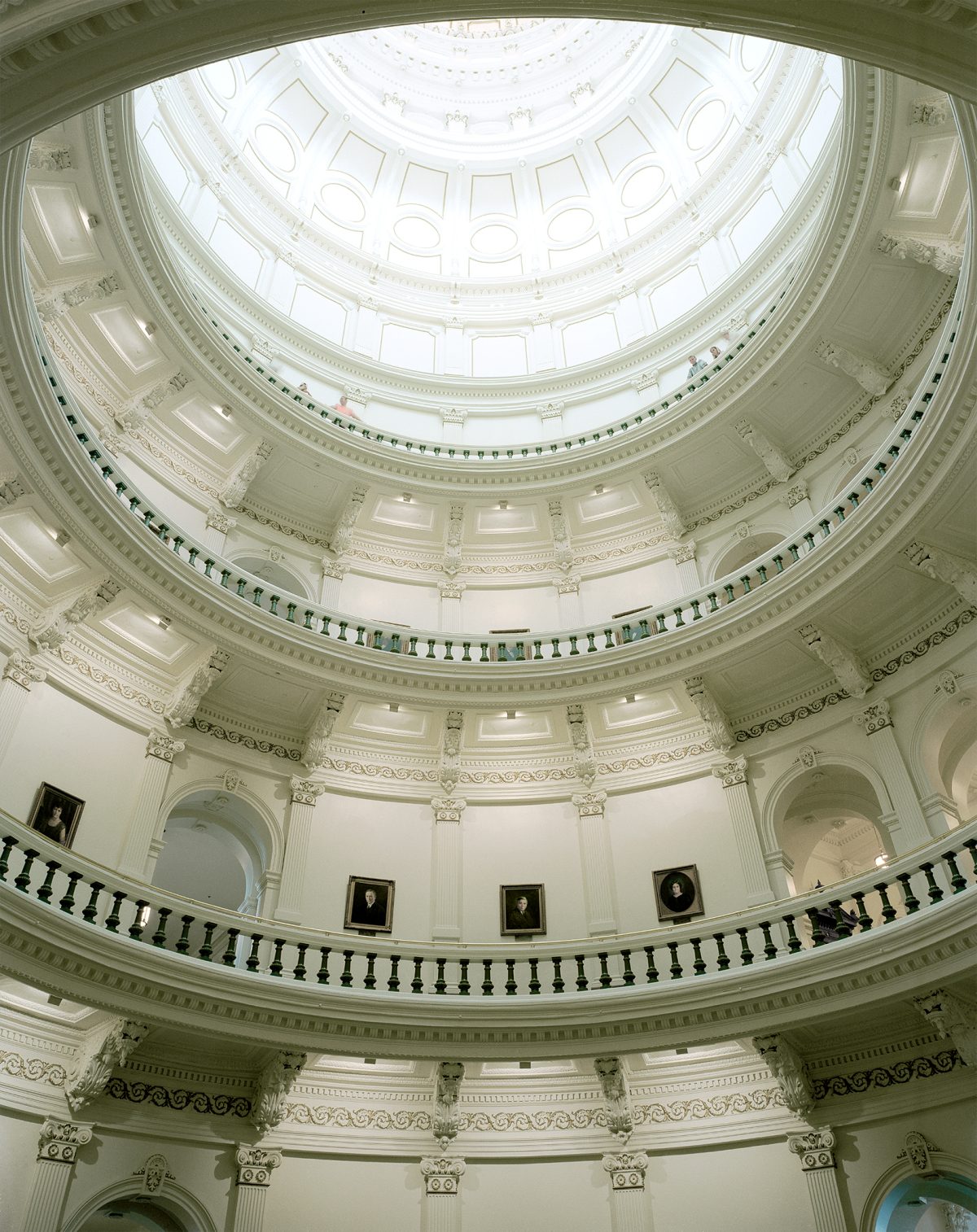 Symmetrical upward view of a grand neoclassical dome interior with concentric circular levels featuring ornate white plaster detailing, Corinthian capitals, and decorative moldings. Multiple tiers of arched openings and balustrades recede toward a central skylit oculus at the apex, with portrait paintings visible on the walls below.