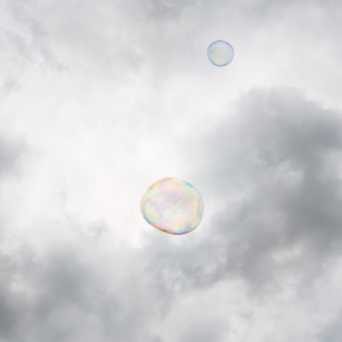 Two iridescent soap bubbles float against a soft, overcast sky with diffused clouds. The larger bubble in the lower center displays subtle rainbow hues across its translucent surface, while a smaller bubble hovers above it, creating a serene, minimalist composition.