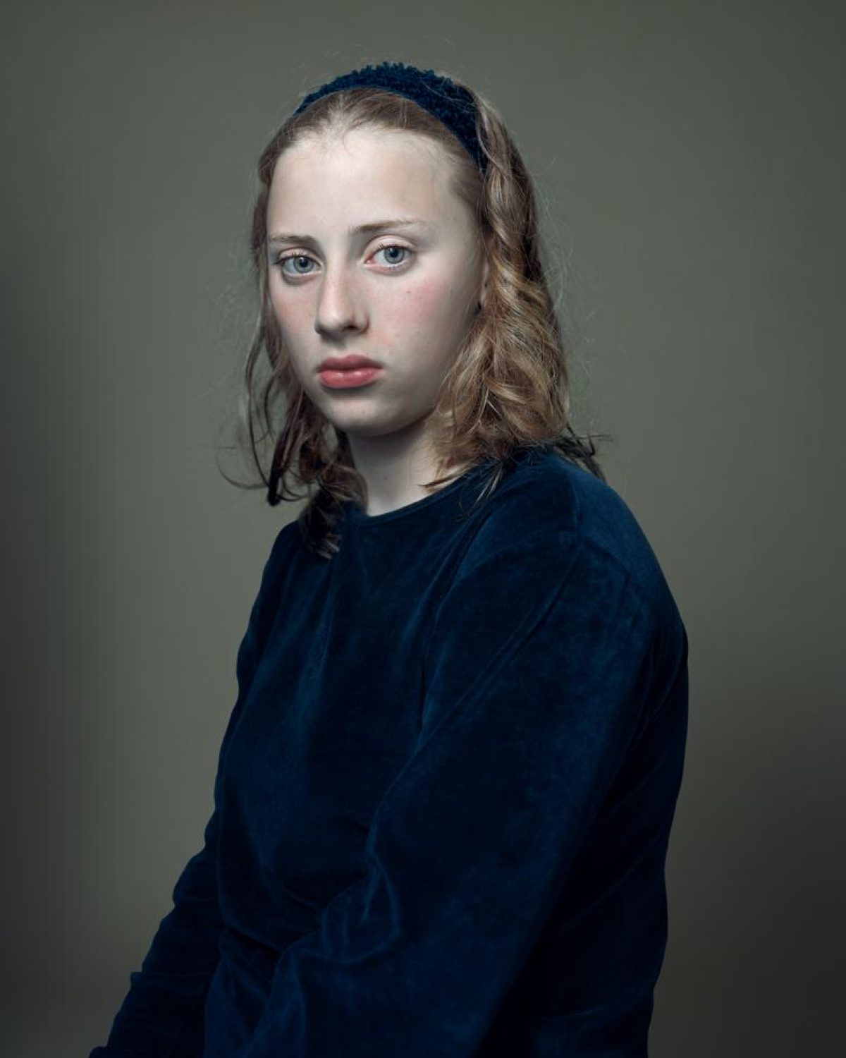 A portrait of a young person with shoulder-length brown hair and pale complexion, wearing a dark blue velvet garment and a blue headband, gazing directly toward the camera against a muted green-gray background.
