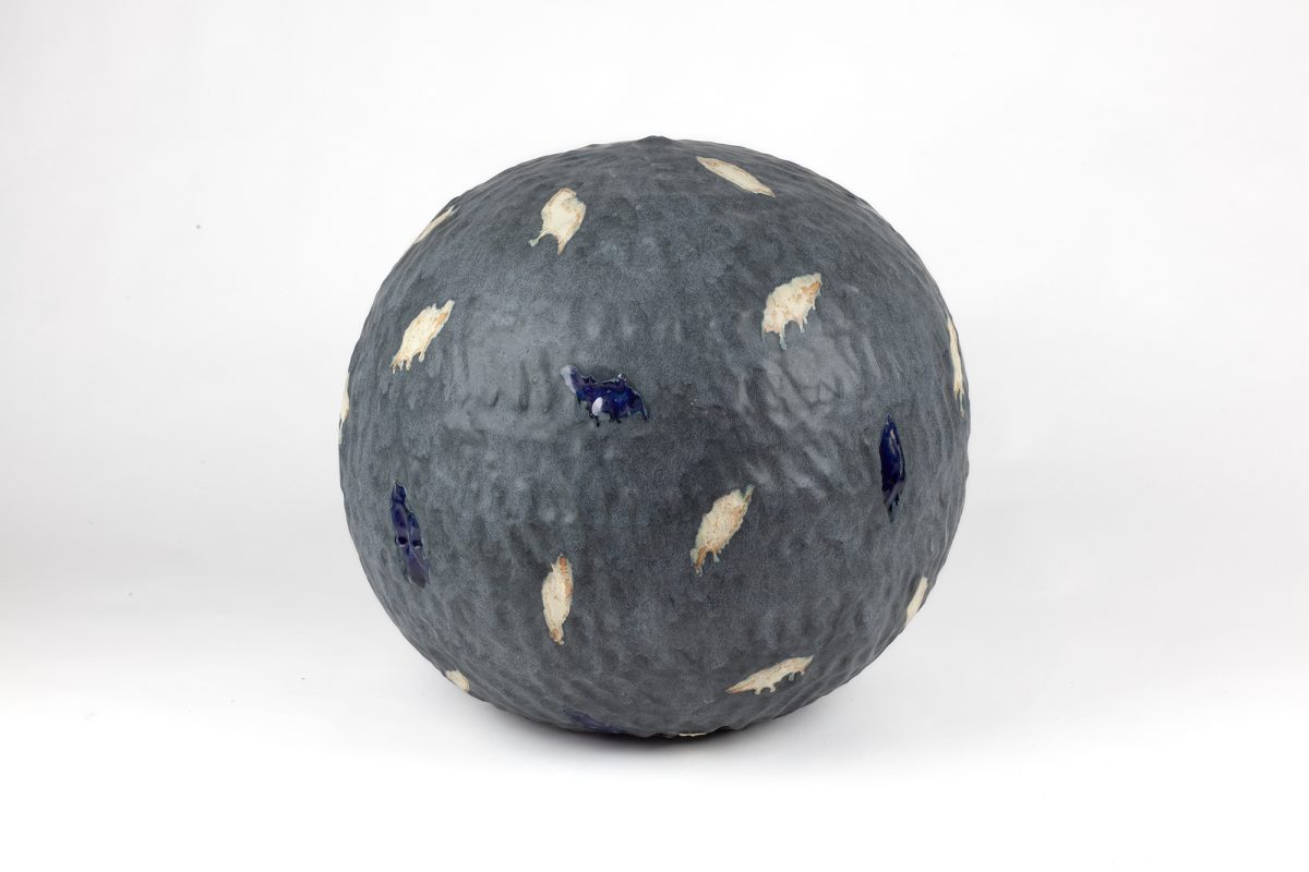 A spherical ceramic form with a textured grey surface scattered with cream-colored and deep blue leaf-shaped appliqués, displayed against a white background.