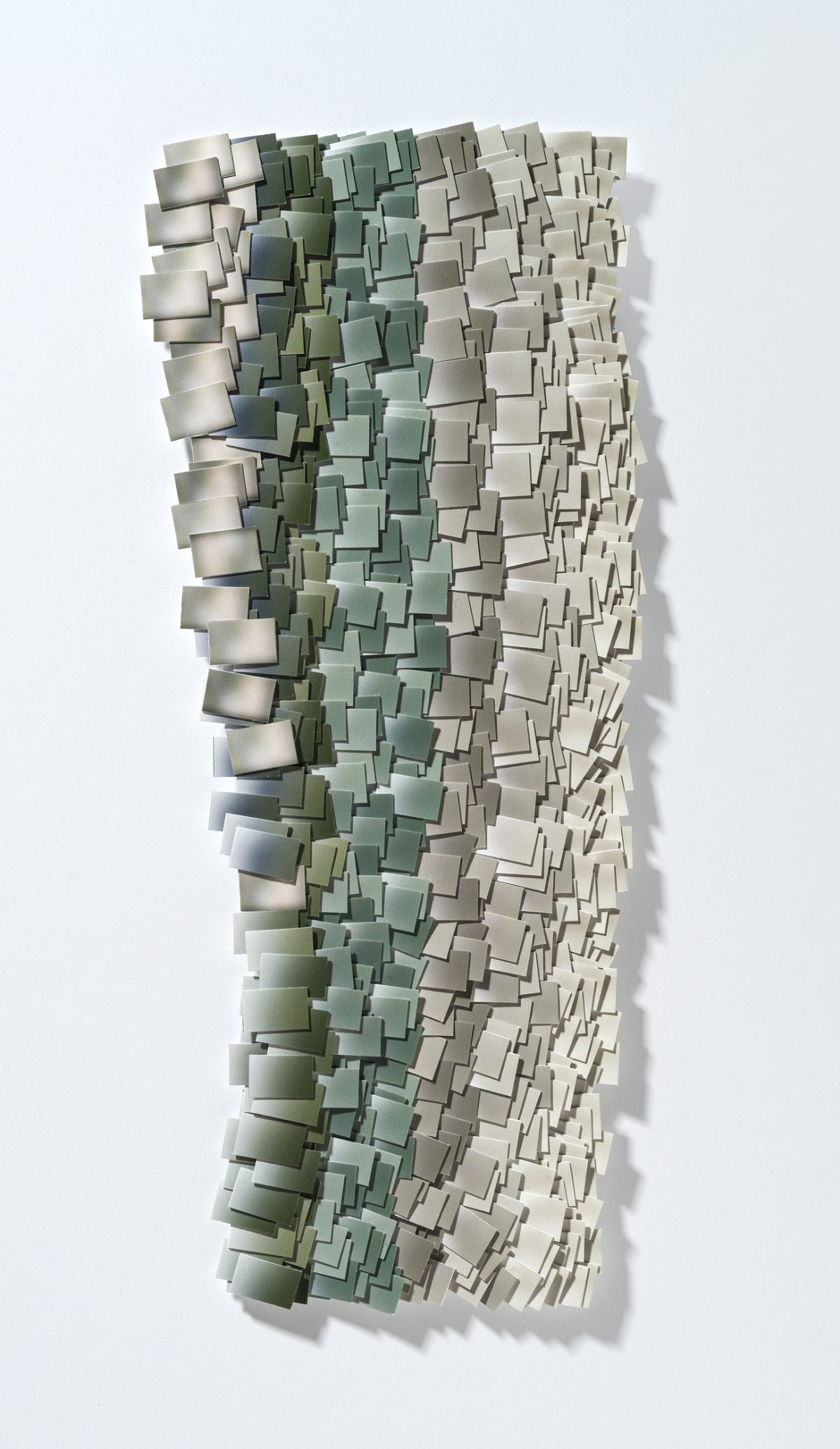 A vertical wall-mounted artwork composed of hundreds of hand-cut and layered rectangular paper fragments in graduated tones of sage green, taupe, and cream, creating a three-dimensional textured surface with pronounced shadows that suggest a pixelated or topographic landscape.
