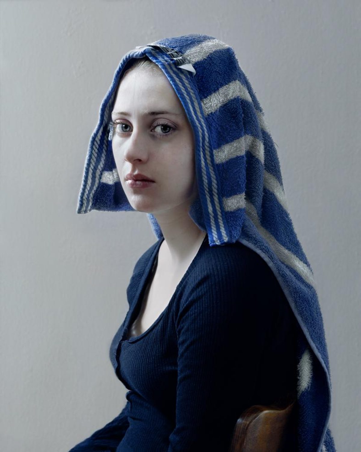 A young woman in profile facing left, wearing a dark ribbed navy garment with a blue and gray striped towel wrapped around her head. She has pale skin, defined facial features, and a composed expression, set against a neutral gray background with warm brown tones visible at the lower right edge.