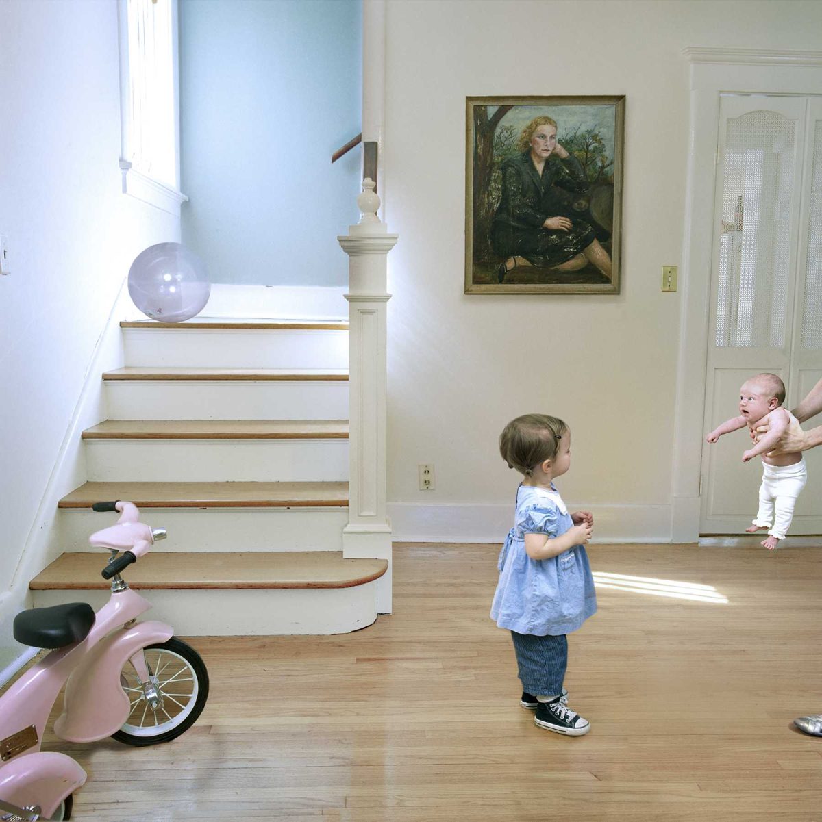A young child in a blue shirt and jeans stands in a bright, sun-filled hallway watching a baby being held by someone on the right. A pink tricycle sits in the foreground on the left, while wooden stairs with a white banister and a framed portrait painting are visible in the background. The scene captures a moment of domestic life in a contemporary home interior.