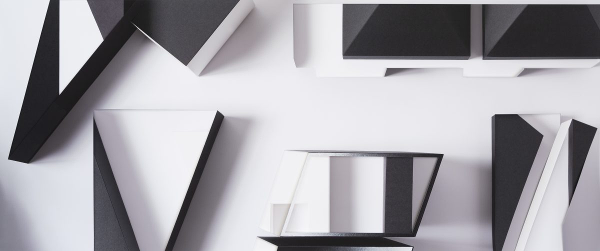 Geometric abstract composition of layered, fragmented forms in black, white, and gray tones arranged across a light background. Sharp angular shapes and rectangular planes overlap and cast shadows, creating depth and visual tension through a deconstructed, collage-like arrangement.