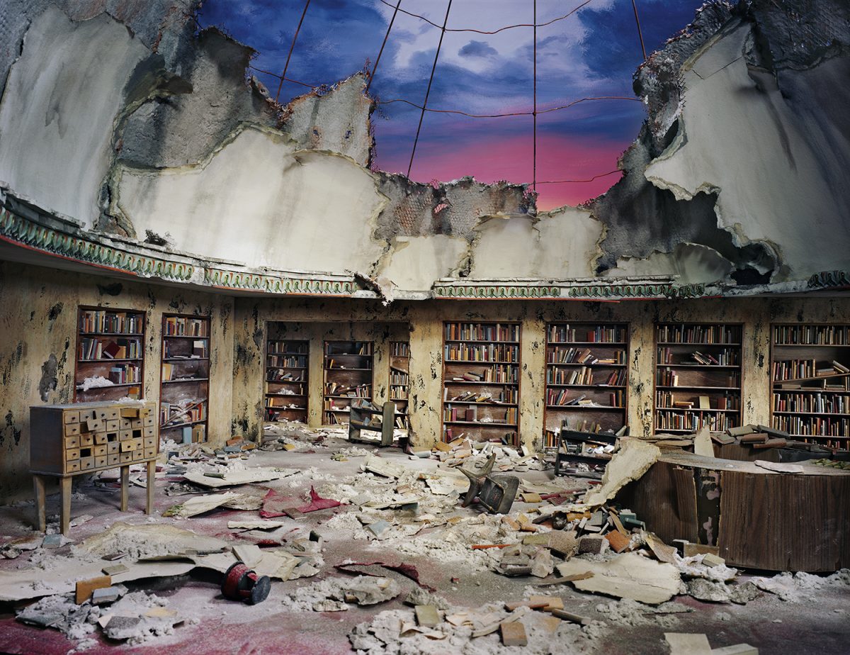 A devastated library interior with rows of bookshelves lining crumbling walls, their spines still visible among scattered debris and rubble covering the floor. The ceiling is partially destroyed, framing a dramatic twilight sky in shades of blue and pink above the scene of decay.
