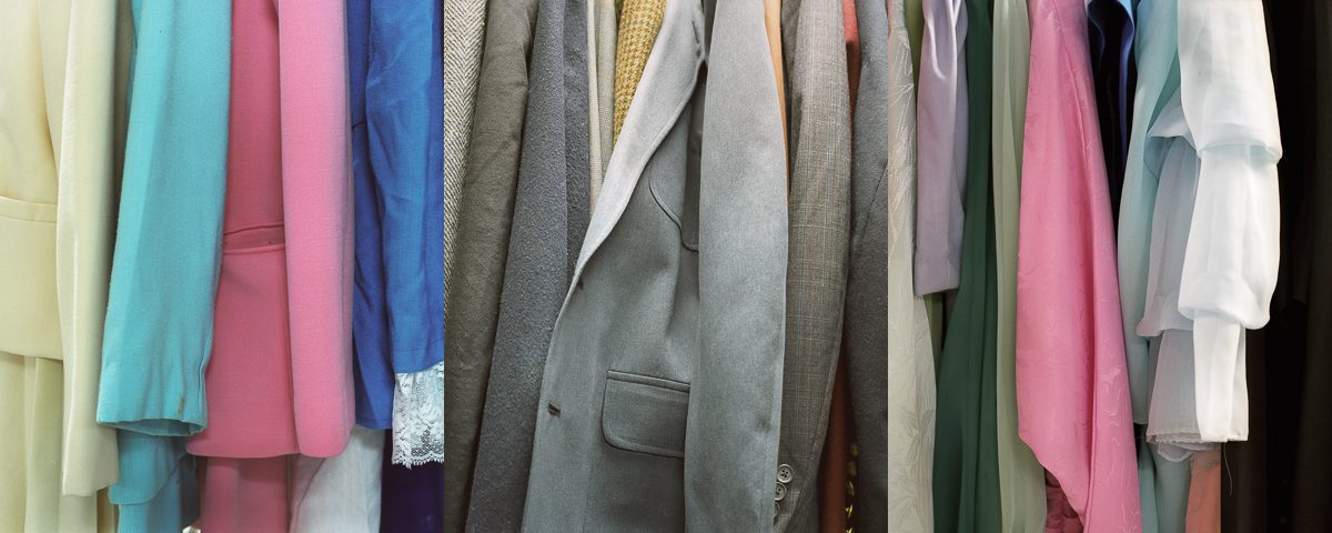 A densely packed row of hanging garments in varied colors including pastels, neutrals, and jewel tones, displayed against a neutral background. The clothing appears layered and overlapping, showcasing different fabrics and textures in shades of pink, blue, gray, green, and white.