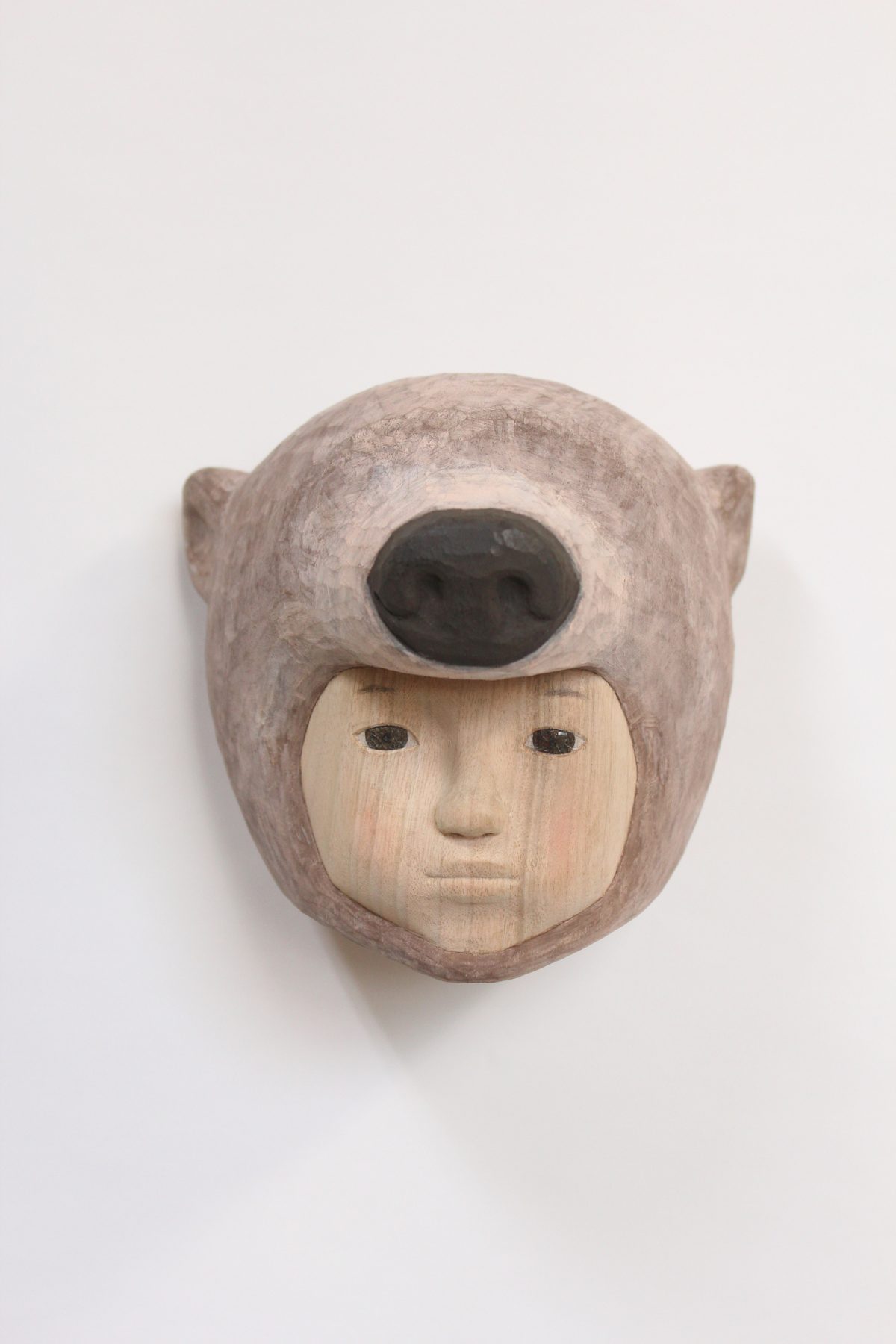 Carved wooden mask depicting a human face with closed eyes and neutral expression, framed by a rounded animal head with pointed ears and a large dark oval opening where the eyes would be. The lighter wood face contrasts with the darker weathered tones of the surrounding animal form.