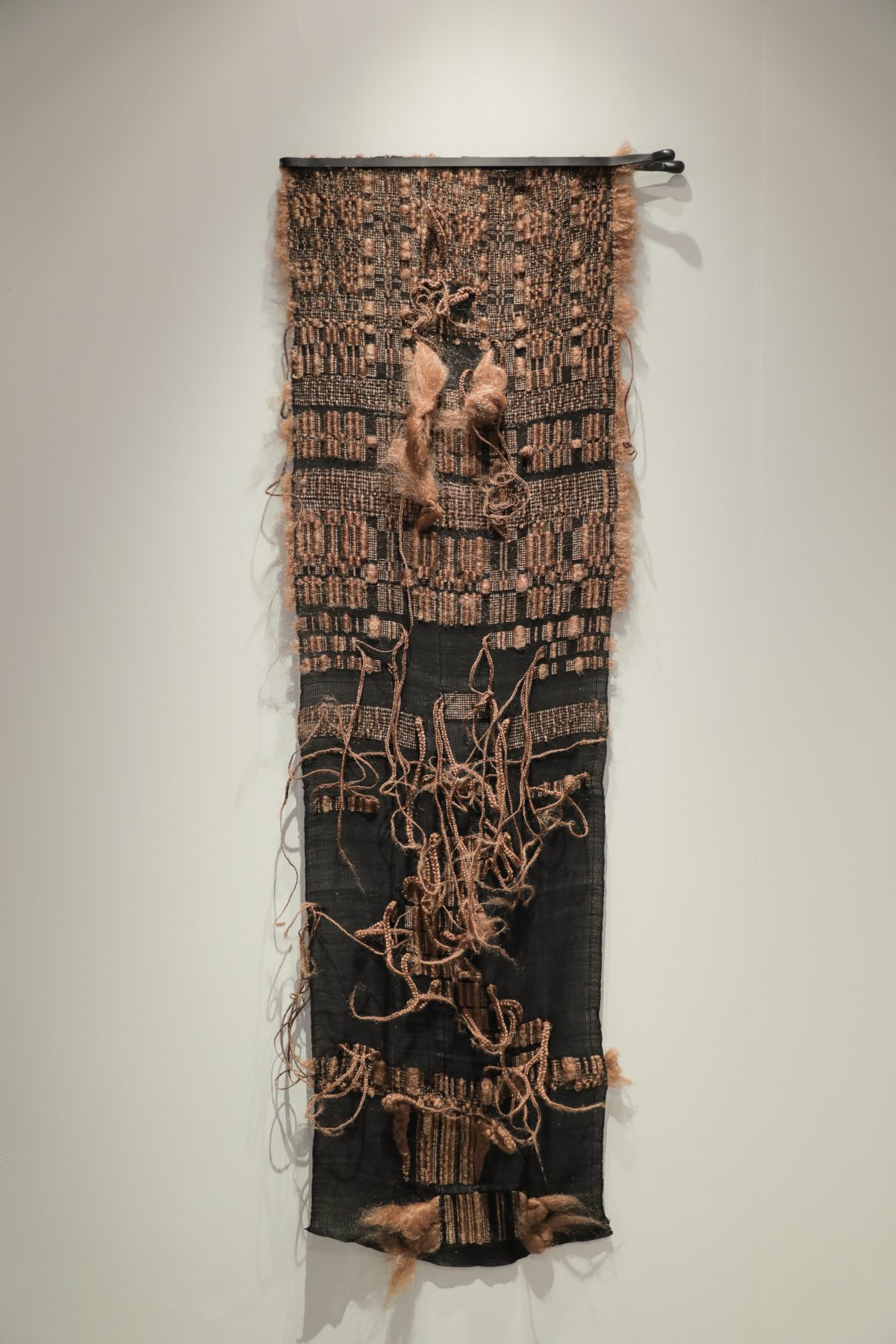 A vertical textile wall hanging combining woven cotton with synthetic hair elements. The upper portion features a tan and black woven pattern with figurative forms, while the lower section is predominantly black with looping strands of blonde hair creating organic, gestural marks across the surface.