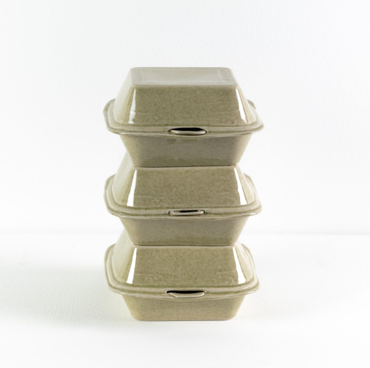 Three stacked porcelain takeout containers with hinged lids in pale celadon green, arranged vertically to form a tower-like sculptural form against a white background.