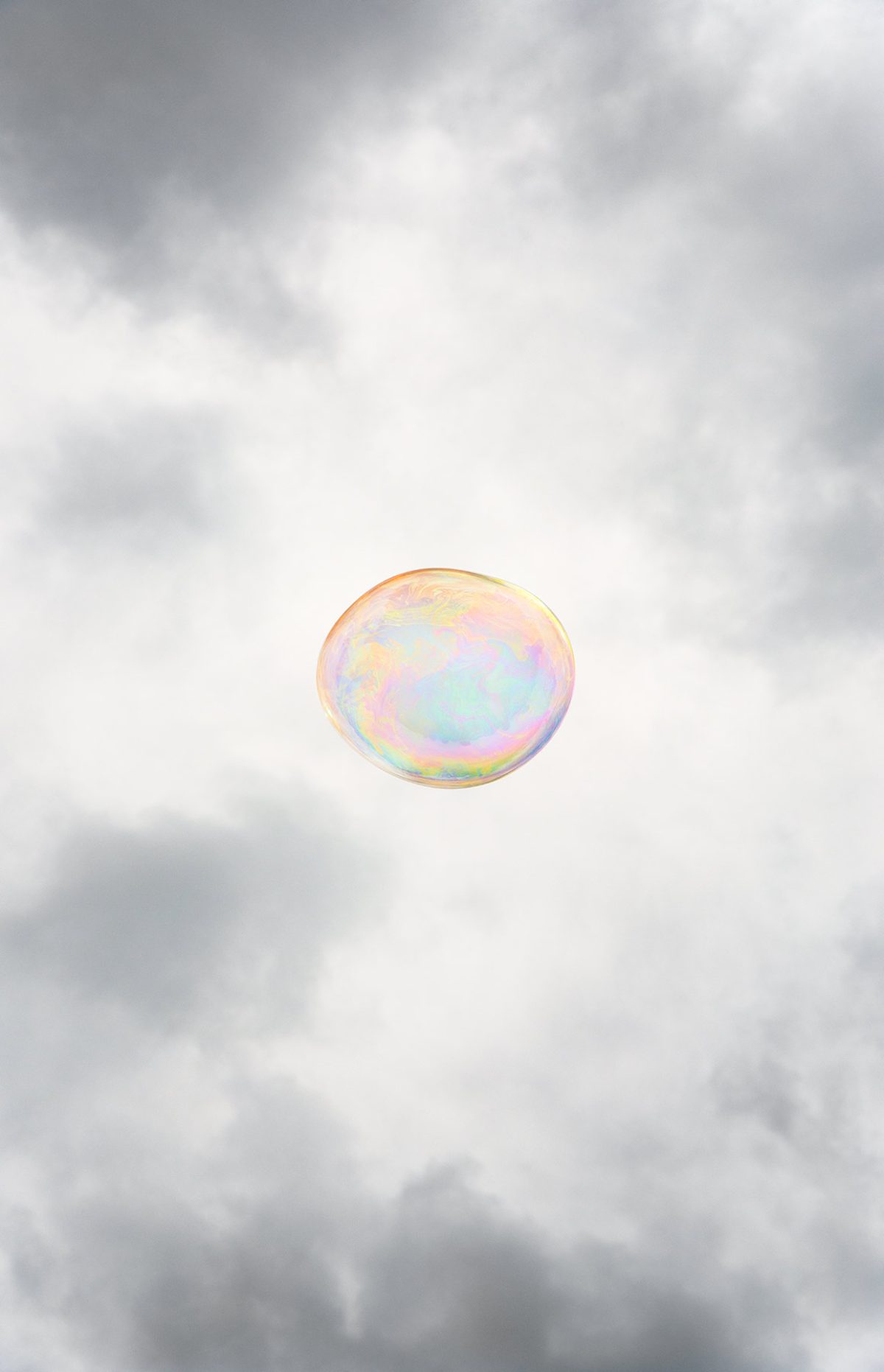A single iridescent soap bubble floats against a cloudy gray sky, its translucent surface displaying soft pastel hues of pink, yellow, and blue that shimmer across its spherical form.