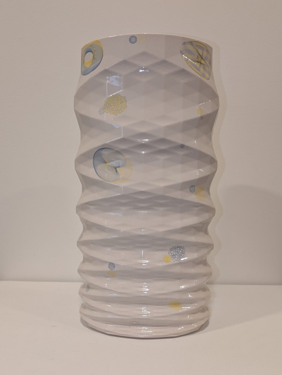 A white porcelain vessel with undulating, organic sides features abstract motifs in soft blue and pale yellow throughout—circular forms, linear patterns, and gestural marks that evoke scientific or natural imagery. The glazed surface catches light across its textured, rhythmically rippled form.