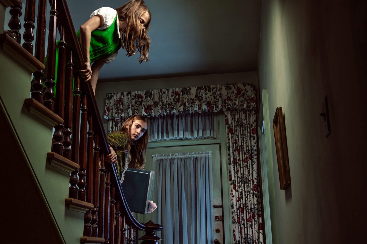 Two figures positioned on a wooden staircase in a mint-green interior with floral curtains and vintage furnishings. The upper figure wears a bright green garment and leans dramatically over the railing, while the lower figure peeks around the banister holding a dark object. Dramatic lighting and Dutch-angle perspective create a surreal, theatrical composition.