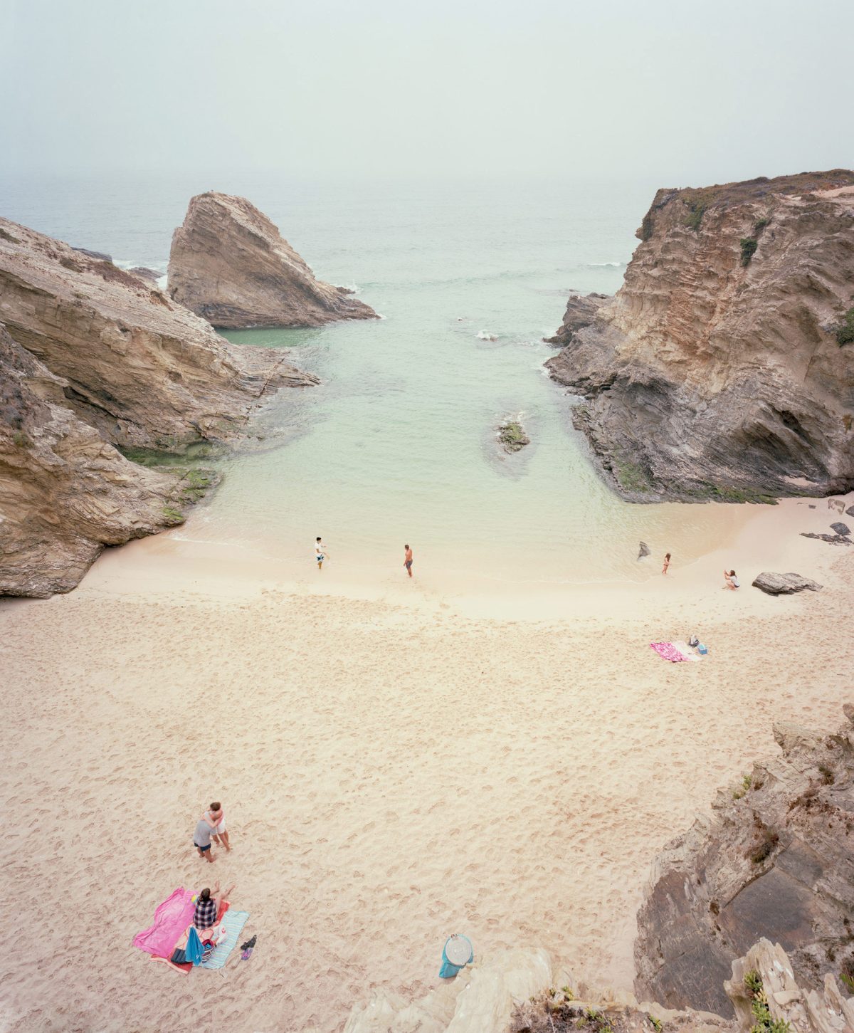 Aerial view of a secluded cove with pale turquoise water framed by dramatic, striated limestone cliffs. A small sandy beach is dotted with scattered beachgoers and colorful beach items, creating a serene summer scene rendered in soft, naturalistic tones.