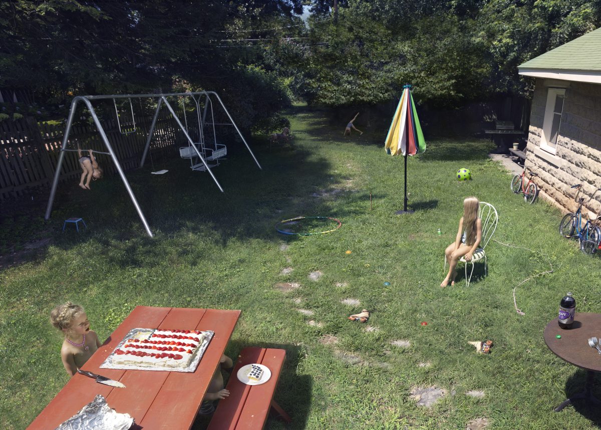 A backyard summer scene with children engaged in various activities: one child sits at a picnic table with a decorated cake, another sits on a chair in the middle distance, and a third swings on a metal swing set. Bicycles lean against a stone wall near a house, a colorful umbrella stands in the grass, and toys are scattered throughout the sunny lawn bordered by trees.