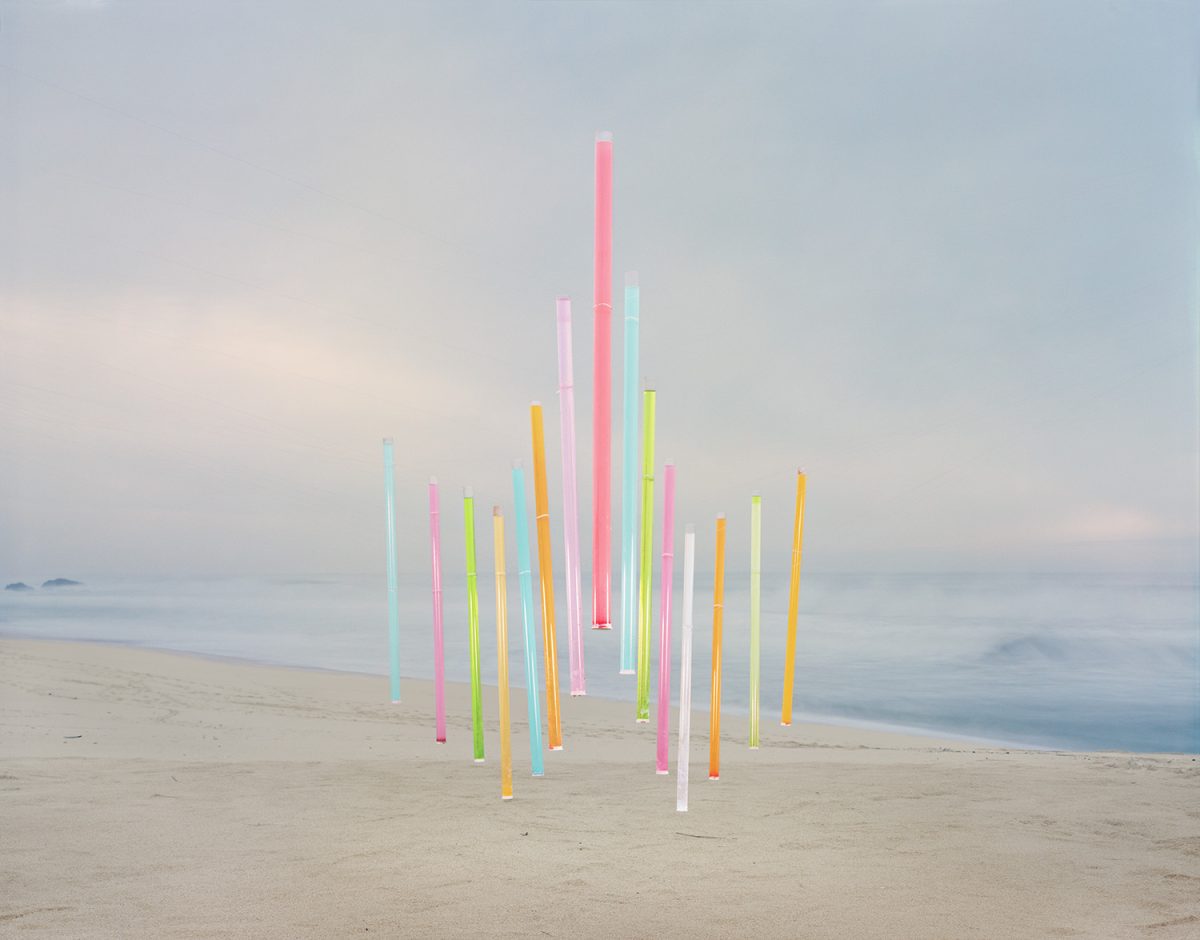 Vertical neon-colored tubes in pink, yellow, cyan, lime green, and white arranged in a clustered formation on a misty beach, with soft gray ocean and sky in the background.