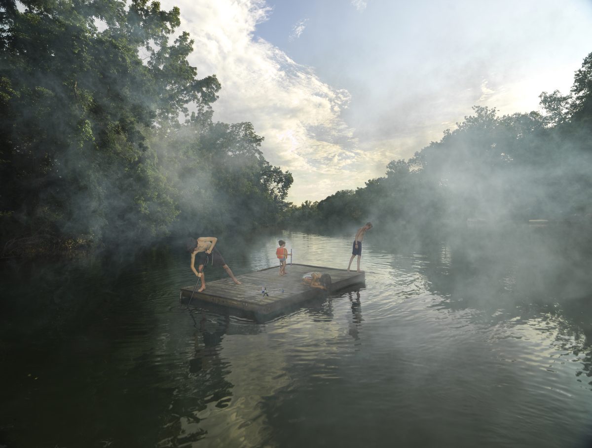 Several figures stand and play on a wooden raft floating in a misty river, surrounded by dense green forest on both banks. Morning or evening light filters through thick fog rising from the water, creating an ethereal atmosphere beneath a clouded sky.