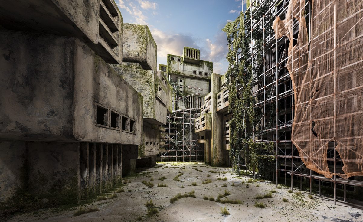 Abandoned concrete buildings with moss-covered facades line a narrow courtyard overgrown with sparse vegetation and sandy ground. Metal scaffolding and safety netting cover the deteriorating structures, while vines climb the weathered walls beneath a partly cloudy sky.