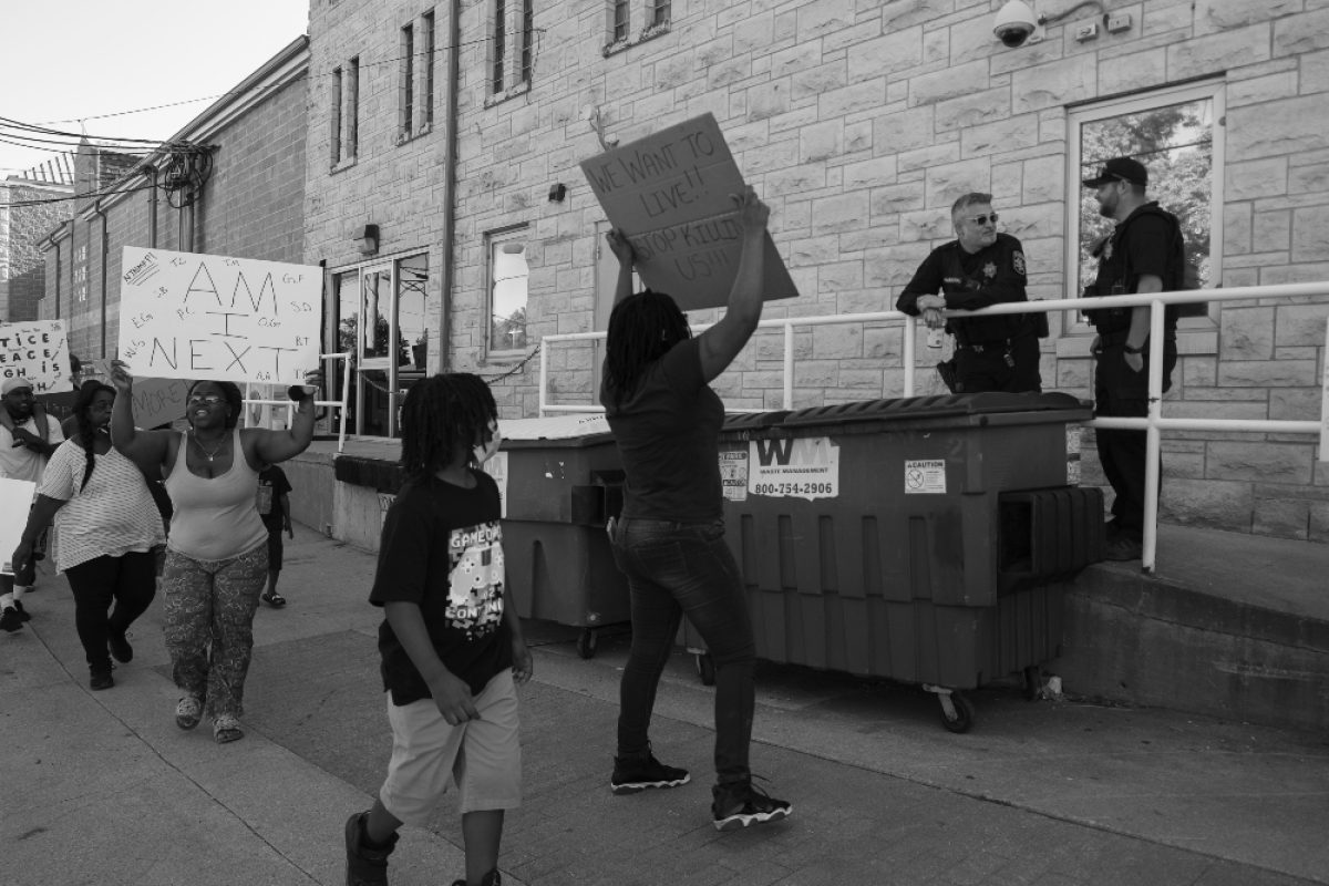 Black and white photograph of a street protest outside a brick building, showing demonstrators holding handmade signs while uniformed officers stand on an elevated platform. The composition captures the tension between the crowd at street level and the authority figures positioned above them on the structure.