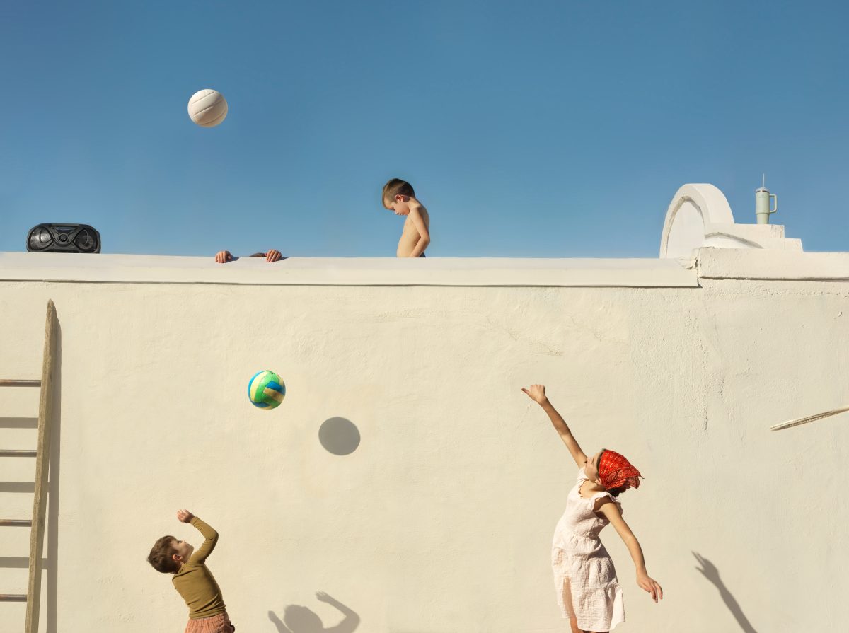 Children play with balls on a stark white concrete wall dividing an upper and lower level against a clear blue sky. A shirtless boy stands on top of the wall watching, while below, two children toss colorful balls upward, their shadows cast sharply against the pale surface. Beach equipment and a white structure are visible at the top edge of the composition.