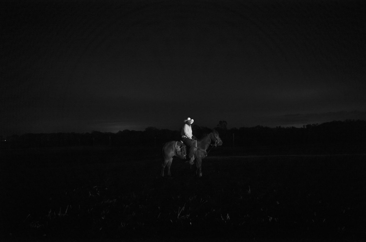 A solitary figure in light clothing sits atop a horse in an expansive, dark landscape at dusk or night. The composition is rendered in high-contrast black and white, with the rider and horse illuminated against a nearly black sky and terrain, creating a stark, isolated presence on the plains.