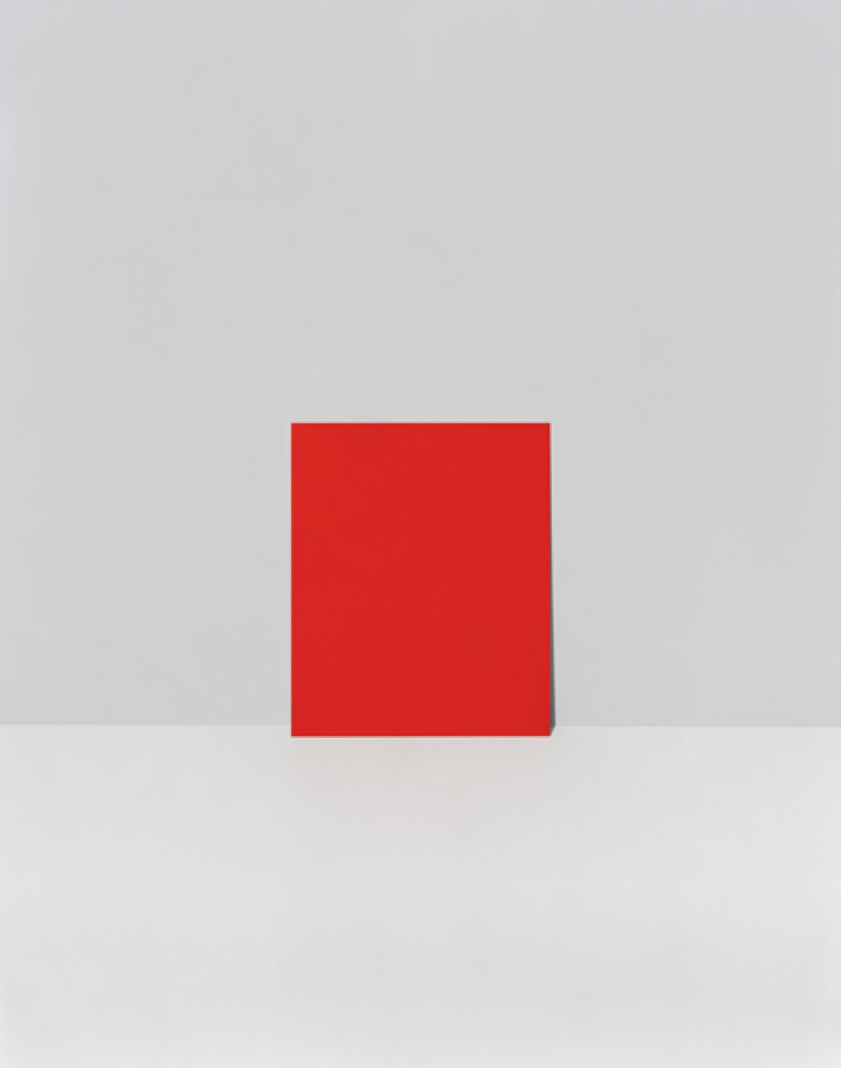 A bright red square positioned centrally against a neutral gray and white background, creating a stark geometric composition with minimal color variation and clean spatial division.