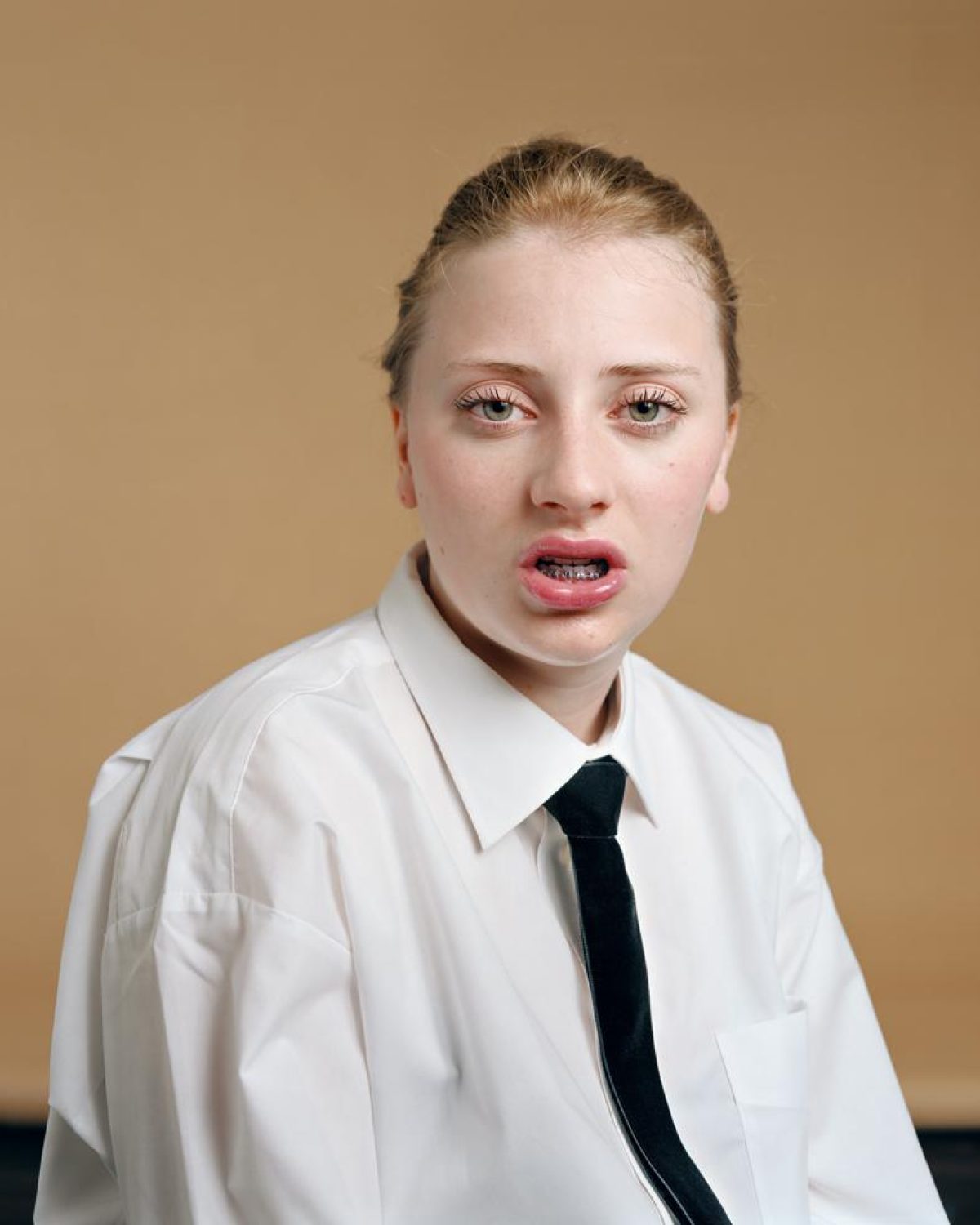 Portrait of a young person with reddish-blonde hair pulled back, wearing a white collared shirt and black tie against a warm tan background. The subject gazes directly at the viewer with a slightly open mouth expression, rendered in vivid color with sharp photographic clarity.