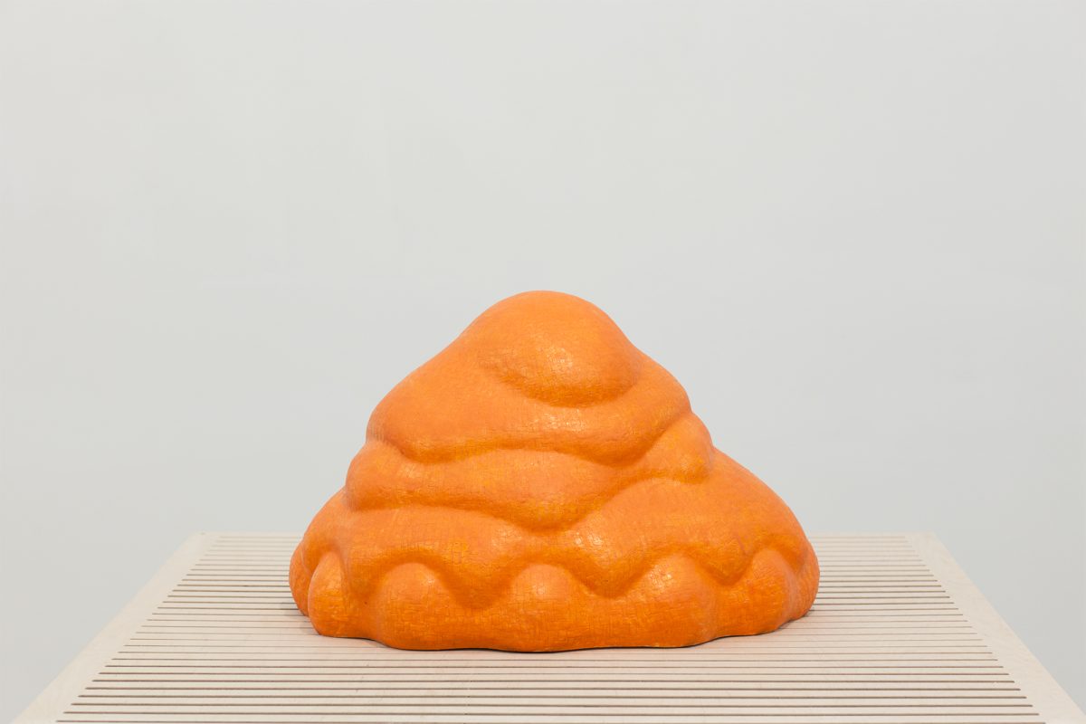 A bright orange glazed ceramic sculpture with an organic, rounded form sits on a striped wooden platform. The bulbous shape features layered, undulating surfaces that suggest both natural and abstract qualities against a neutral gray background.