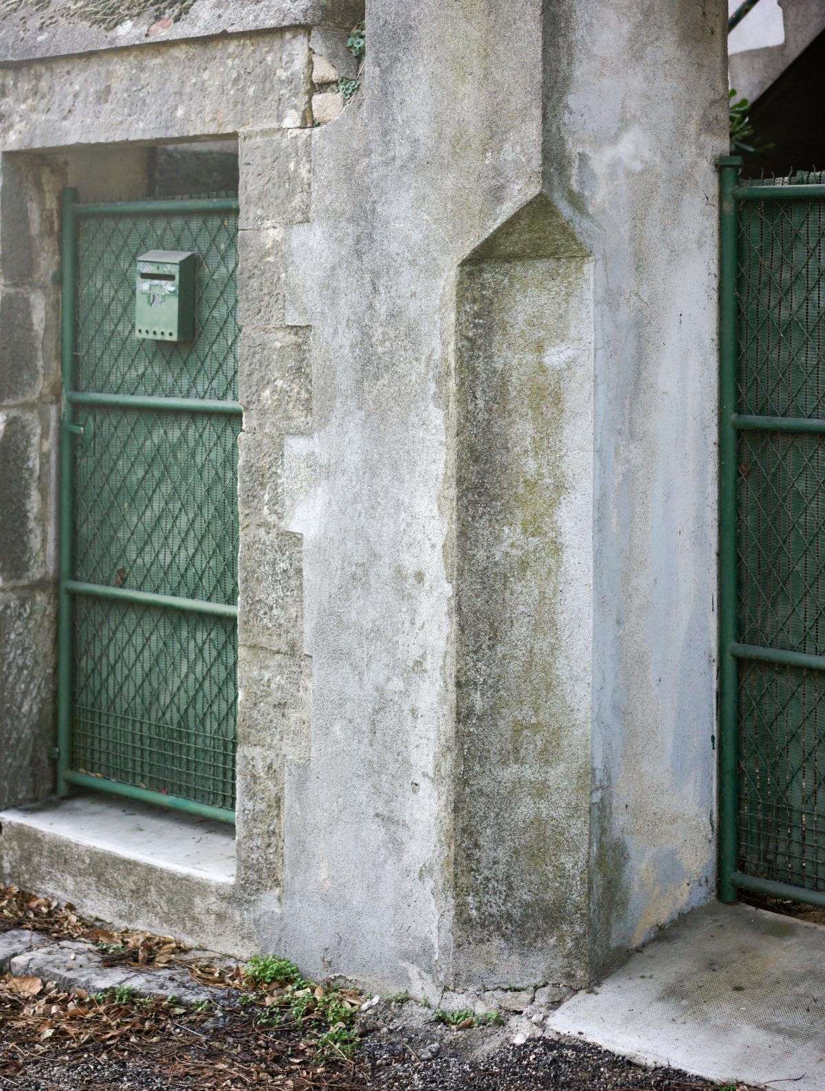 Weathered concrete pillars and walls marked by moss and lichen staining frame a narrow passageway lined with green metal gates and a mailbox, capturing the texture and decay of an industrial or utilitarian structure.